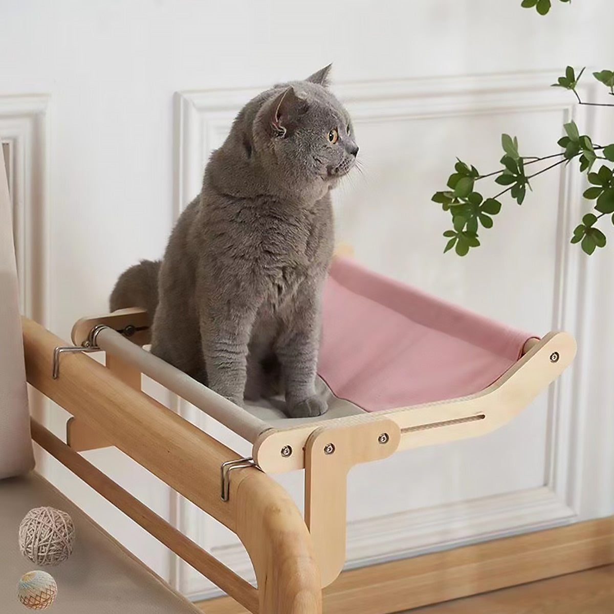 A gray cat sitting on a pink adjustable hanging cat hammock bed placed by a window with plants in the background.
